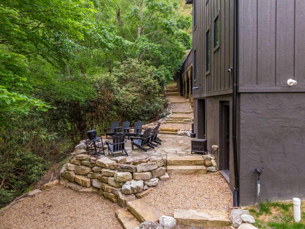 A stone patio surrounded by lush greenery, featuring a set of Adirondack chairs arranged for relaxing or socializing. A pathway leads through the natural garden setting, suggesting seamless access to nature and outdoor enjoyment.