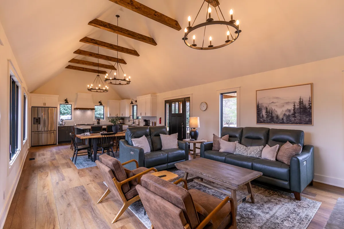 A warm, open-plan great room featuring plush sofas, a large dining table, and seamless sightlines into a modern kitchen. The space is illuminated by abundant natural light, accented by decorative lighting fixtures and tasteful artwork. The cozy atmosphere is centered around the inviting seating and fireplace, ideal for gathering.