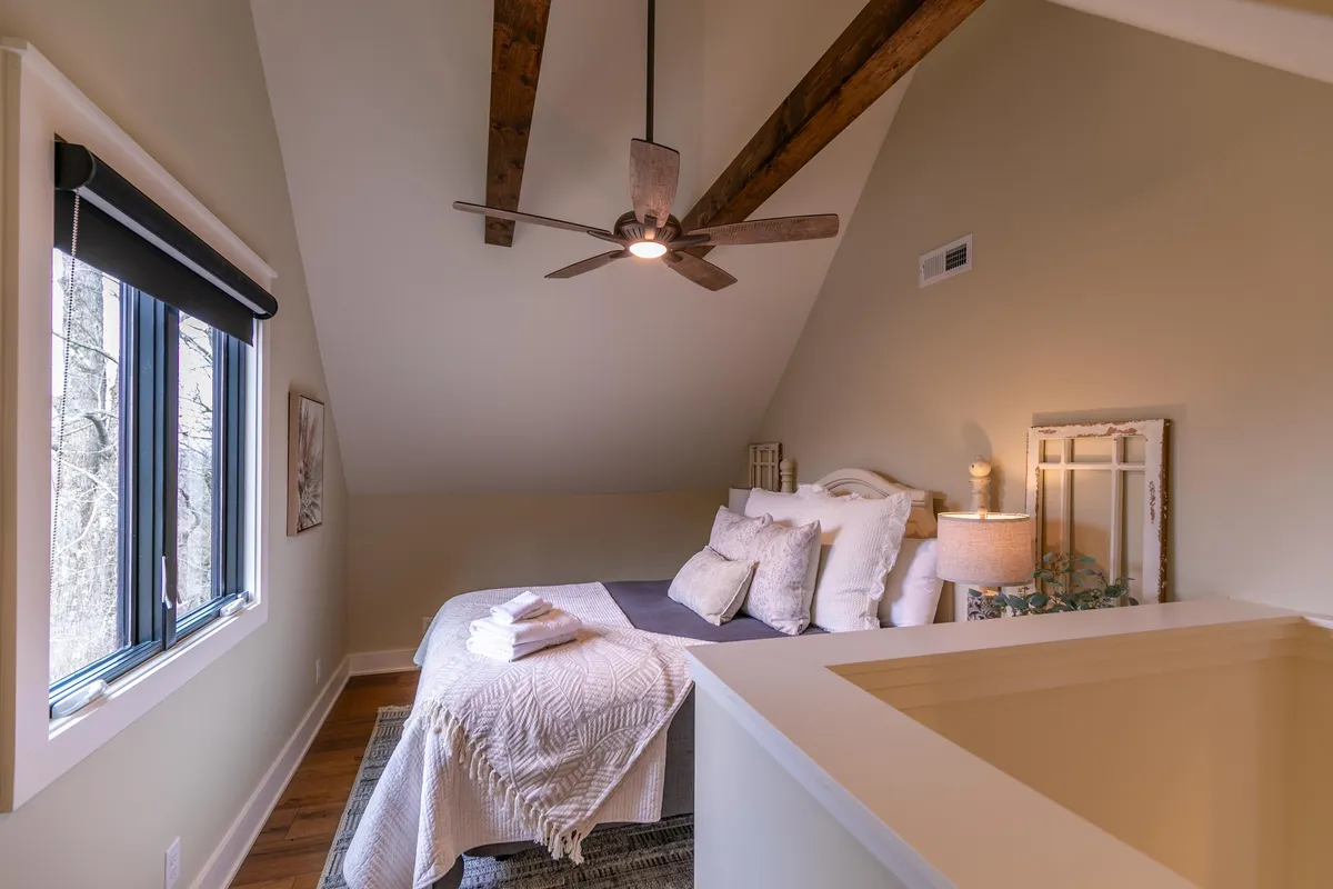 A comfortable Queen bedroom with a plush bed, ceiling fan, bedside lamp, window, and decorative artwork. The soft, inviting atmosphere is illuminated by gentle natural light, offering a peaceful spot for solo guests or couples to recharge.