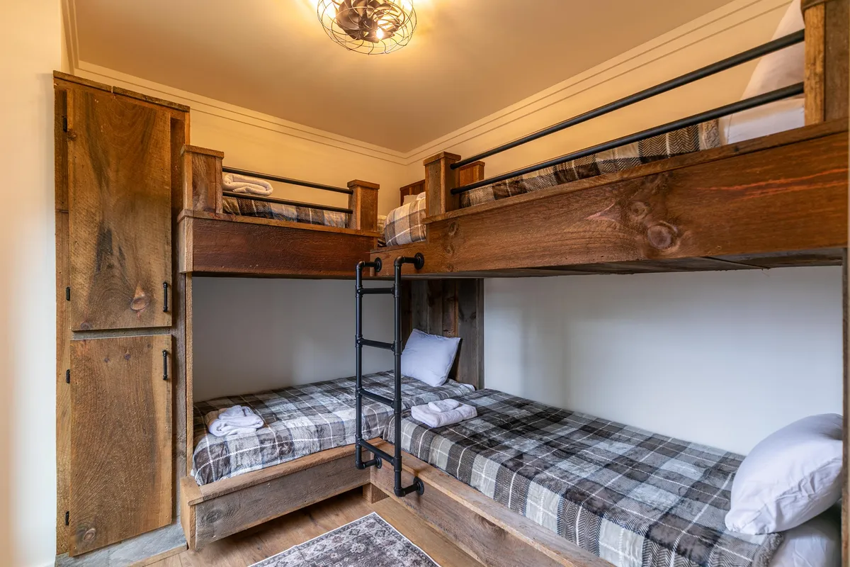 A warm and inviting bunkroom featuring two sets of built-in bunk beds with plaid bedding and wooden cabinetry. The space is cozy and playful, designed to foster memorable sleepovers and fun for younger guests or groups of siblings.