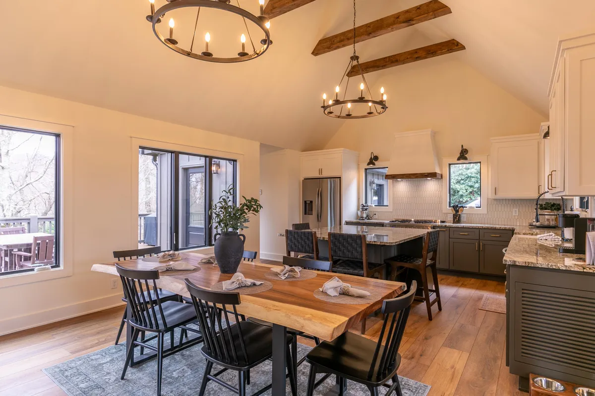A bright, inviting dining area anchored by a spacious dining table under elegant chandeliers. The kitchen island, complete with granite countertops, is visible in the background, and large windows flood the space with natural light and offer glimpses outside.
