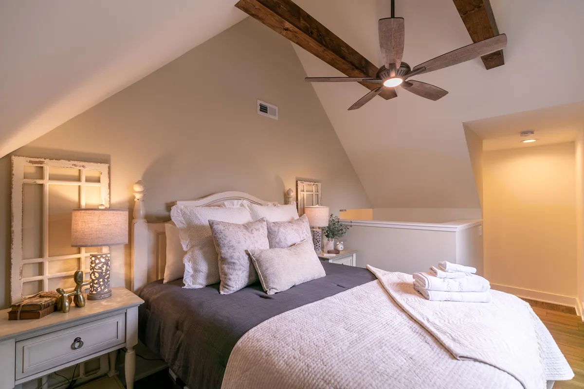 A serene King suite with a plush bed adorned with decorative pillows, nightstands with lamps on either side, and a ceiling fan above. The space is softly lit by natural light streaming in from the window, creating an atmosphere perfect for relaxing, reading, or unwinding after a day in the mountains.