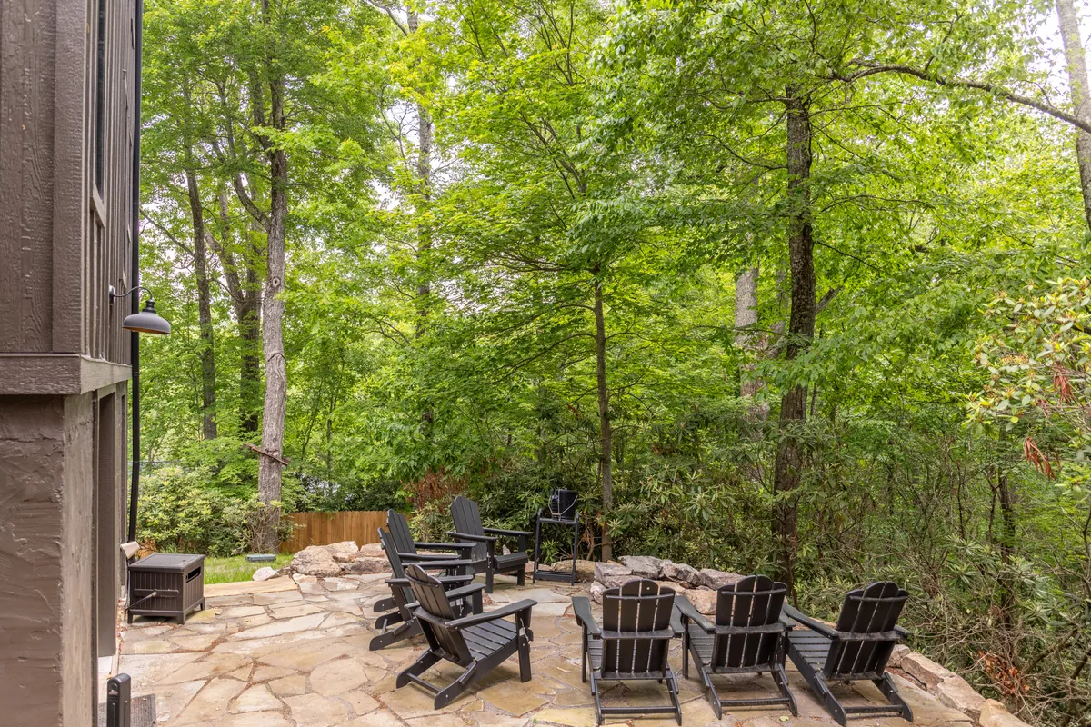 A serene stone patio surrounded by trees and set with black Adirondack chairs. The setting is tranquil and encourages relaxation, providing a space for group conversations or quiet moments, immersed in nature.