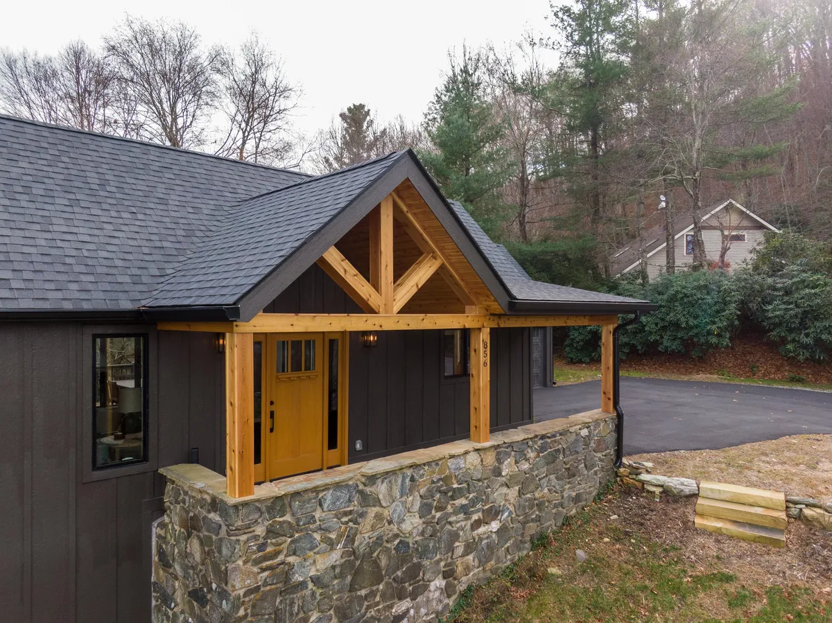 An inviting exterior porch with a yellow front door, wooden structure, and stone wall welcomes guests. The porch appears perfect for gatherings, with space for friends or family to relax and enjoy the outdoors. The surrounding nature hints at a peaceful setting, ideal for creating memories.