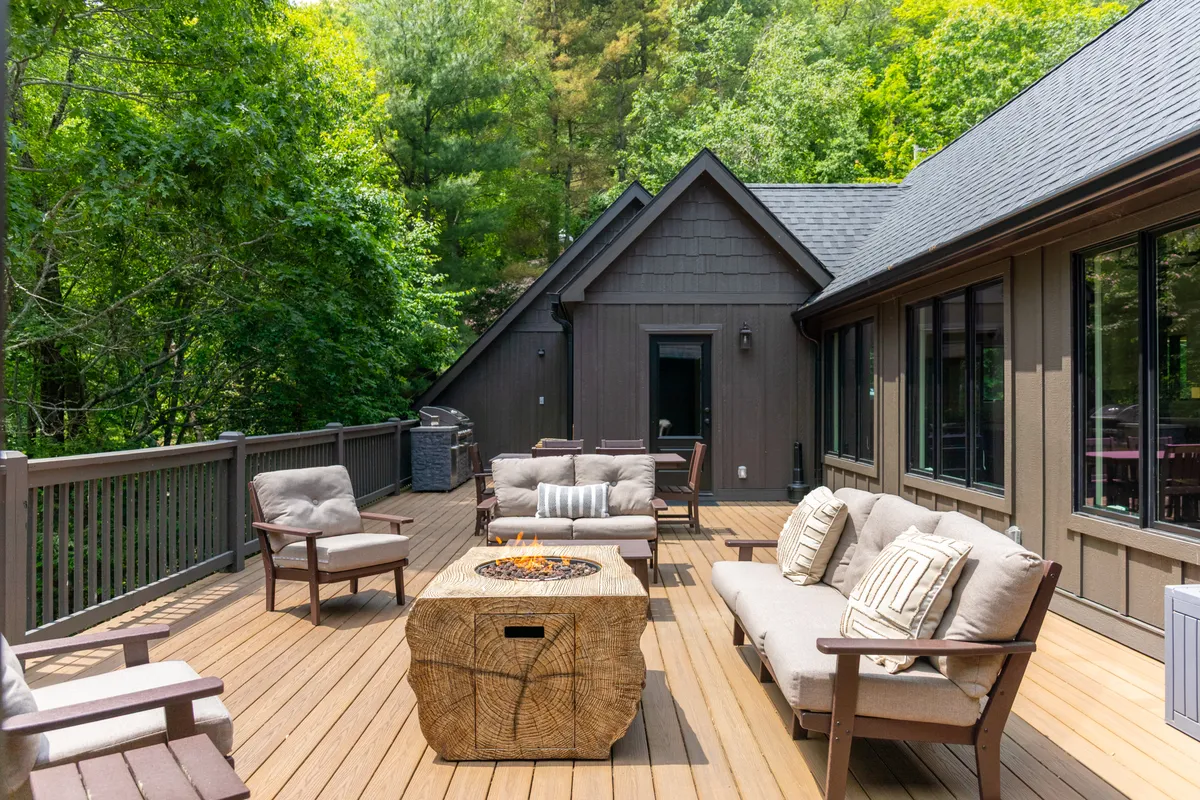 An outdoor fire pit set on a wooden deck encircled by Adirondack chairs, with trees and sunset hues in the background. It conveys a cozy, communal atmosphere ideal for unwinding, roasting marshmallows, or chatting under the stars.